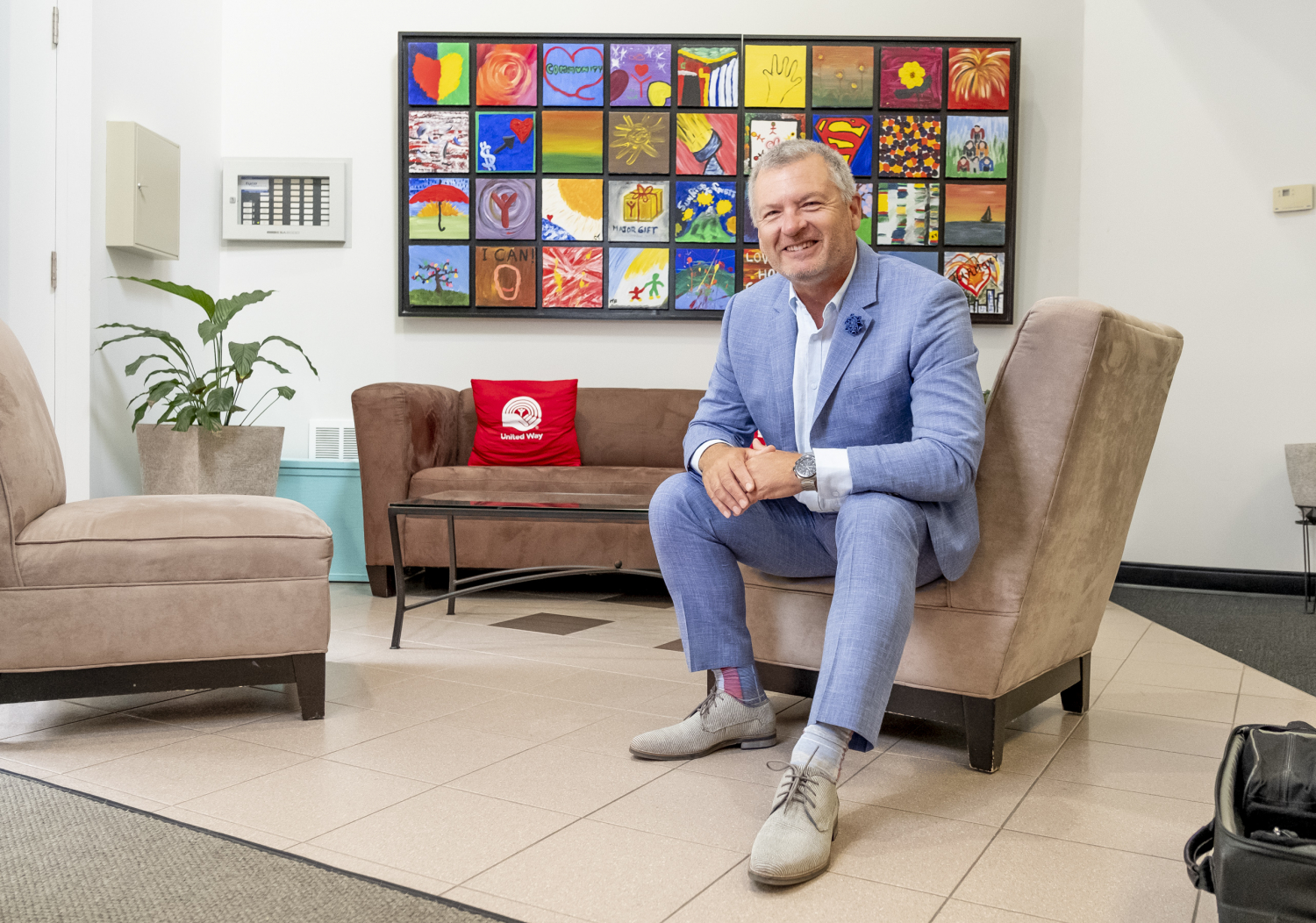 A smiling, white haired man in a blue linen suit sitting in front of a mural of painted panels